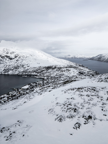 Aerial view of snow covered mountains surrounding a deep blue fjord and a small coastal village under an overcast sky Ersfjord Troms Norway. Aerial view of snow covered mountains surrounding a deep blue fjord and a small coastal village under an overcast sky Ersfjord Troms Norway.