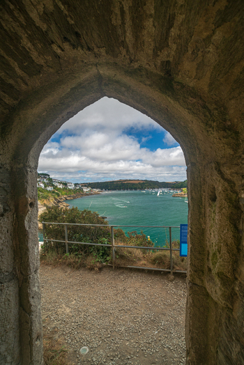 View of the Fowey River and the town of Fowey seen through a historic stone archway with boats moored in the water under a blue sky in Fowey Cornwall United Kingdom. View of the Fowey River and the town of Fowey seen through a historic stone archway with boats moored in the water under a blue sky in Fowey Cornwall United Kingdom.