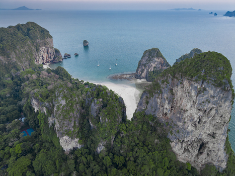 Aerial view of dramatic limestone cliffs cloaked in lush greenery meet a pristine white sand beach and turquoise waters Krabi Thailand.