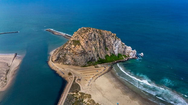 Aerial view of Morro Rock volcanic plug surrounded by the blue Pacific Ocean and sandy beaches with a parking area nearby in Morro Bay California United States.