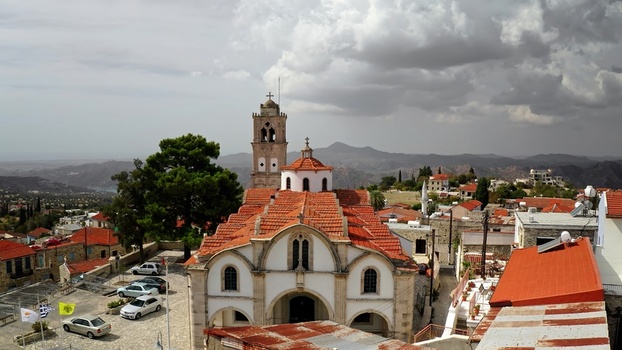 Aerial view of a historic church with a red tiled roof and bell tower situated in a village surrounded by rolling hills and scattered buildings in Cyprus. Aerial view of a historic church with a red tiled roof and bell tower situated in a village surrounded by rolling hills and scattered buildings in Cyprus.