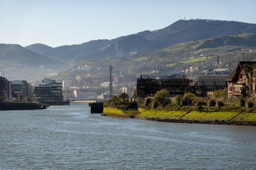 View of the Nervion River flowing past industrial ruins and modern architecture with green mountains in the background Greater Bilbao Basque Country Spain. View of the Nervion River flowing past industrial ruins and modern architecture with green mountains in the background Greater Bilbao Basque Country Spain.
