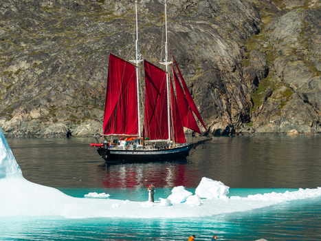 Aerial view of a sailboat with striking red sails navigating near a large iceberg in the icy waters of Greenland. Aerial view of a sailboat with striking red sails navigating near a large iceberg in the icy waters of Greenland.