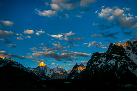 View of the golden summit of Gasherbrum IV mountain peak during sunset with snow covered ridges and scattered clouds in the Karakoram range Skardu Gilgit Pakistan. View of the golden summit of Gasherbrum IV mountain peak during sunset with snow covered ridges and scattered clouds in the Karakoram range Skardu Gilgit Pakistan.