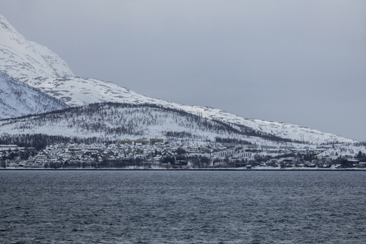 View of a coastal village with traditional houses nestled at the base of snow covered mountains under an overcast sky in Kvaloya Troms Norway. View of a coastal village with traditional houses nestled at the base of snow covered mountains under an overcast sky in Kvaloya Troms Norway.