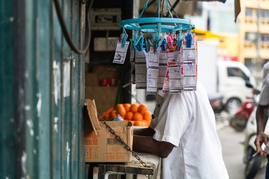 Sri Lanka 15 February 2026 View of a street vendor stall with lottery tickets hanging from a blue plastic rack a man in a white shirt and oranges in the background.