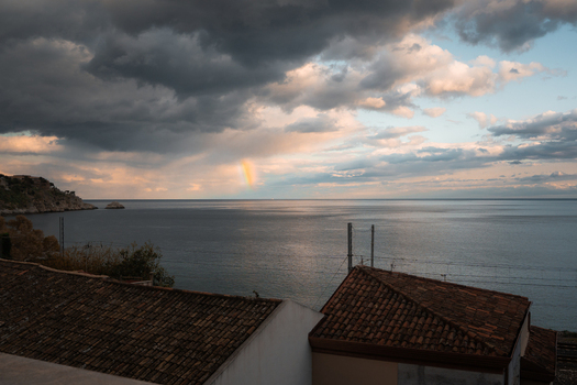 View of a faint rainbow arcing over the tranquil sea under a dramatic cloud streaked sky terracotta rooftops adding warmth to the coastal scene Taormina Sicily Italy.