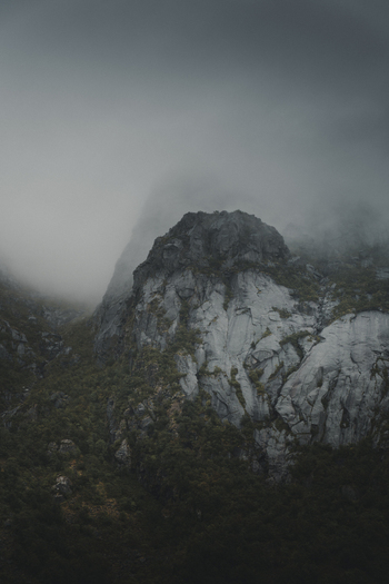 View of the craggy mountain peaks of Lofoten partially shrouded in thick mist and low clouds with dark green vegetation on the slopes in Nordland Norway. View of the craggy mountain peaks of Lofoten partially shrouded in thick mist and low clouds with dark green vegetation on the slopes in Nordland Norway.