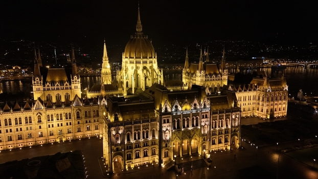 Aerial view of the illuminated Hungarian Parliament Building at night featuring its intricate Gothic Revival architecture and glowing facade along the Danube River in Budapest Hungary.