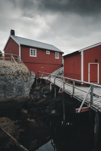 Nusfjord Norway 07 September 2022 View of traditional red wooden cabins on stilts connected by a wooden walkway in the historic fishing village of Nusfjord under a cloudy sky. Nusfjord Norway 07 September 2022 View of traditional red wooden cabins on stilts connected by a wooden walkway in the historic fishing village of Nusfjord under a cloudy sky.