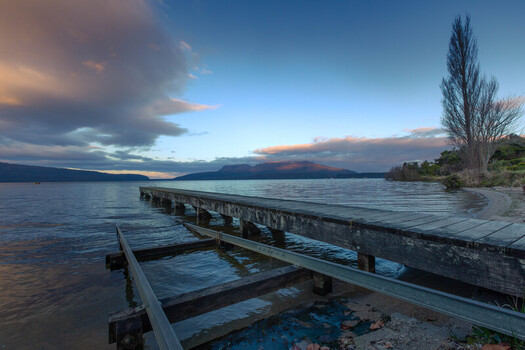 View of a wooden jetty extending into the calm waters of Lake Tarawera at sunset with distant mountains and soft clouds in the sky in Rotorua Bay of Plenty Region New Zealand. View of a wooden jetty extending into the calm waters of Lake Tarawera at sunset with distant mountains and soft clouds in the sky in Rotorua Bay of Plenty Region New Zealand.