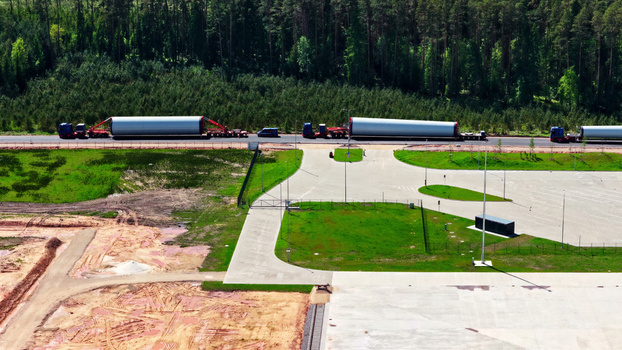 Aerial view of trucks transporting large wind turbine blades along a road bordered by a dense forest and a construction site in Latvia. Aerial view of trucks transporting large wind turbine blades along a road bordered by a dense forest and a construction site in Latvia.