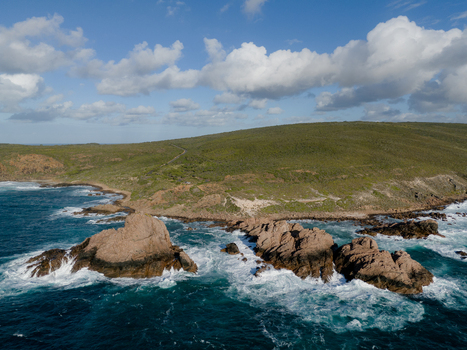 Aerial view of Sugarloaf Rock surrounded by crashing waves and turquoise ocean with green coastal hills under a blue sky in Naturaliste Western Australia Australia.