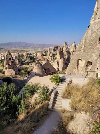 View of Cappadocia rock formations and fairy chimneys with stone stairs leading down a path and ancient cave dwellings under a clear blue sky in Turkiye. View of Cappadocia rock formations and fairy chimneys with stone stairs leading down a path and ancient cave dwellings under a clear blue sky in Turkiye.