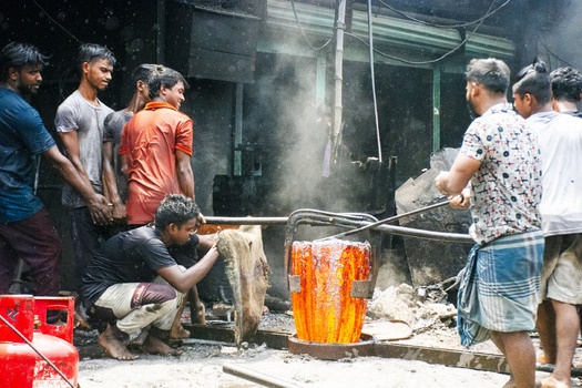 Keraniganj Bangladesh 28 August 2022 View of workers pouring molten metal from a glowing crucible in a shipyard foundry with smoke and dust.