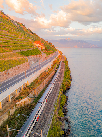 Aerial view of a train traveling along the tracks beside Lake Geneva with terraced vineyards on the hillside and a road above during golden hour in Bourg en Lavaux Vaud Switzerland. Aerial view of a train traveling along the tracks beside Lake Geneva with terraced vineyards on the hillside and a road above during golden hour in Bourg en Lavaux Vaud Switzerland.