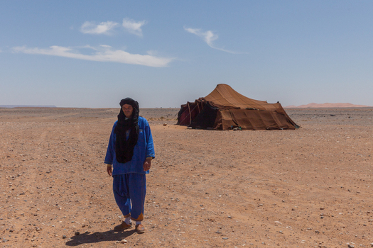View of a man in traditional blue clothing and a black turban standing in the desert near a large brown tent under a clear blue sky in Morocco.