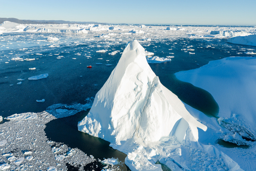 Aerial view of Ilulissat glacier featuring a massive white iceberg floating in the deep blue sea with scattered ice fragments and a small red boat nearby in Greenland. Aerial view of Ilulissat glacier featuring a massive white iceberg floating in the deep blue sea with scattered ice fragments and a small red boat nearby in Greenland.