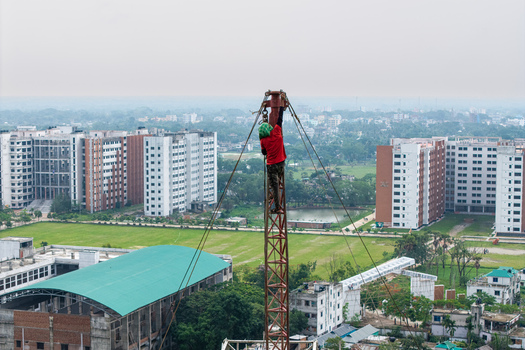 Trishal Upazila Bangladesh 13 May 2026 Aerial view of a worker climbing a tall metal tower overlooking modern university buildings and green fields. Trishal Upazila Bangladesh 13 May 2026 Aerial view of a worker climbing a tall metal tower overlooking modern university buildings and green fields.