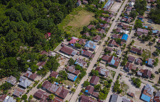 Aerial view of a coastal village with houses featuring metal roofs and dirt roads surrounded by lush tropical vegetation in Raja Ampat Southwest Papua Indonesia.