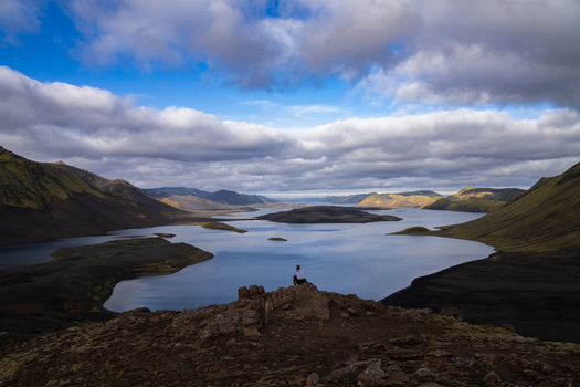 View of a solitary figure perched atop rugged rocks gazing out over the serene lake surrounded by stark majestic mountains under a sky Skaftárhreppur Iceland. View of a solitary figure perched atop rugged rocks gazing out over the serene lake surrounded by stark majestic mountains under a sky Skaftárhreppur Iceland.