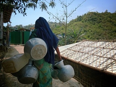 Cox s Bazar Bangladesh 19 July 2026 View of a woman carrying metal water pots through a refugee camp with bamboo shelters and lush green hills in the background. Cox s Bazar Bangladesh 19 July 2026 View of a woman carrying metal water pots through a refugee camp with bamboo shelters and lush green hills in the background.