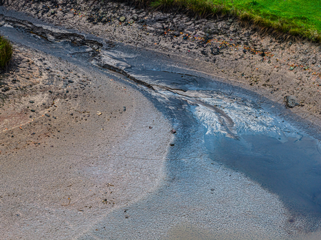 Aerial view of a dried up riverbed with dark wet mud and sediment surrounded by patches of green grass in Latvia.
