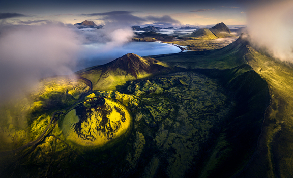 Aerial view of a moss covered volcanic crater and a serene lake surrounded by rugged mountains and low clouds in Iceland.