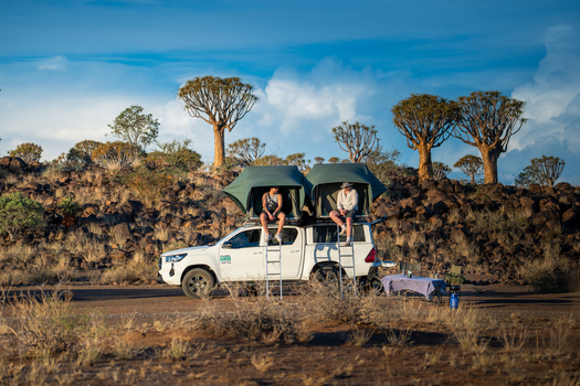 Aussenkehr Namibia 06 March 2026 View of two people perched atop a rooftop tent on a white vehicle framed by the stark sculptural forms of quiver trees against a backdrop of the arid landscape. Aussenkehr Namibia 06 March 2026 View of two people perched atop a rooftop tent on a white vehicle framed by the stark sculptural forms of quiver trees against a backdrop of the arid landscape.