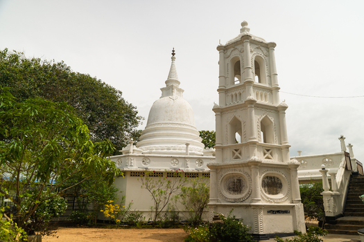 View of a traditional Buddhist temple featuring a white stupa and an ornate bell tower surrounded by lush greenery under an overcast sky in Dodanduwa Southern Province Sri Lanka. View of a traditional Buddhist temple featuring a white stupa and an ornate bell tower surrounded by lush greenery under an overcast sky in Dodanduwa Southern Province Sri Lanka.