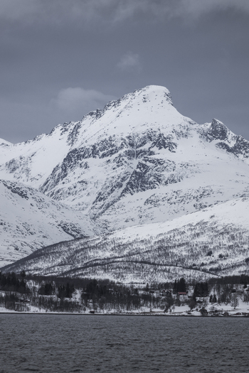 View of a majestic snow covered mountain peak rising above a dark fjord with a small coastal settlement at its base under a cloudy sky in Tromso Troms Norway.