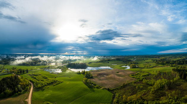 Aerial view of a lush green rural landscape with a small lake rolling meadows patches of forest and a dirt road under a dramatic cloudy sky in Latvia. Aerial view of a lush green rural landscape with a small lake rolling meadows patches of forest and a dirt road under a dramatic cloudy sky in Latvia.