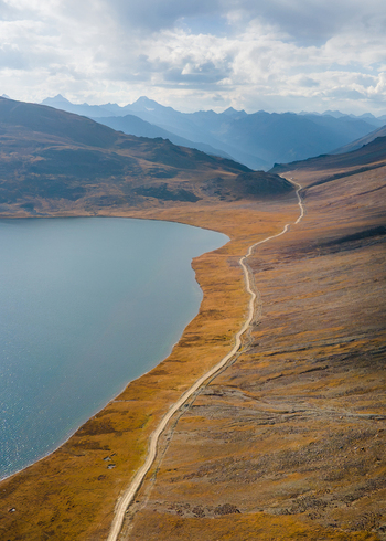 Aerial view of Sheosar Lake with a winding dirt road along the golden shoreline surrounded by vast mountain ranges under a cloudy sky in Skardu Gilgit Pakistan.
