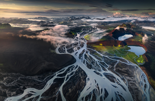 Aerial view of the intricate patterns of braided glacial rivers flowing through volcanic landscapes with craters and misty mountains under a golden sunset Iceland.