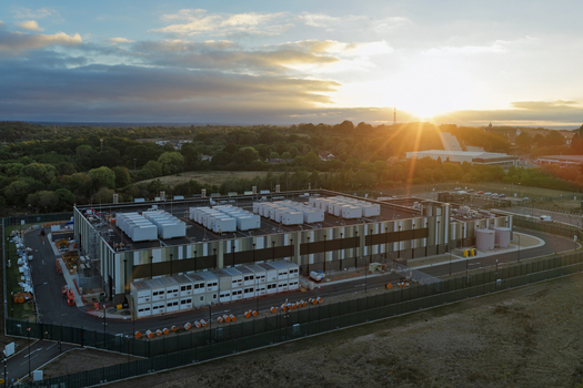 Aerial view of a modern data center facility with white cooling units on the roof surrounded by a perimeter fence and lush greenery under a golden sunset sky in the United Kingdom. Aerial view of a modern data center facility with white cooling units on the roof surrounded by a perimeter fence and lush greenery under a golden sunset sky in the United Kingdom.
