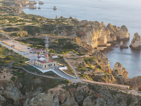 Aerial view of Ponta da Piedade Lighthouse perched on rugged cliffs overlooking the Atlantic Ocean with wooden walkways and golden hour light in Lagos Faro District Portugal.