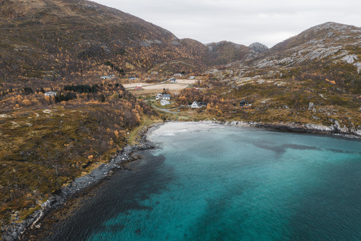 Aerial view of the Senja coastline featuring a small bay with turquoise water surrounding rocky hills with autumn foliage and scattered houses in Senja Troms Norway.