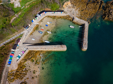 Aerial view of Port Racine with small boats moored in clear turquoise harbor water surrounded by stone breakwaters and colorful sheds along the rocky coast in La Hague Normandie France.