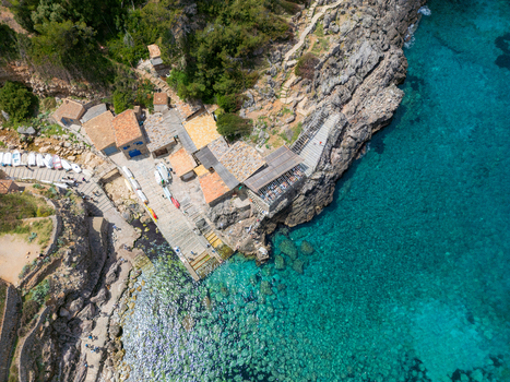 Aerial view of Cala Deia featuring stone buildings a small pier and crystal clear turquoise water along the rocky coastline in Spain. Aerial view of Cala Deia featuring stone buildings a small pier and crystal clear turquoise water along the rocky coastline in Spain.