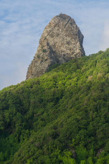 View of the iconic Morro do Pico rock formation rising above lush tropical vegetation and green forest under a soft blue sky in Fernando de Noronha State of Pernambuco Brazil. View of the iconic Morro do Pico rock formation rising above lush tropical vegetation and green forest under a soft blue sky in Fernando de Noronha State of Pernambuco Brazil.