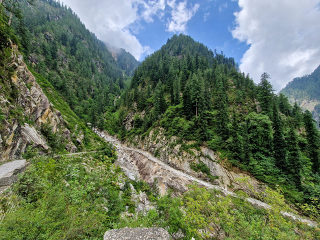 View of the rugged mountain valley with a rocky stream bed dense pine forests covering the steep slopes and a cloudy blue sky in Malana Himachal Pradesh India.