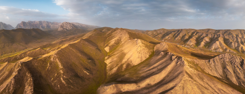 Aerial view of the rugged mountain landscape near Kol Suu featuring rolling golden hills dramatic rocky peaks and a vast sky in At Bashi District Naryn Region Kyrgyzstan.