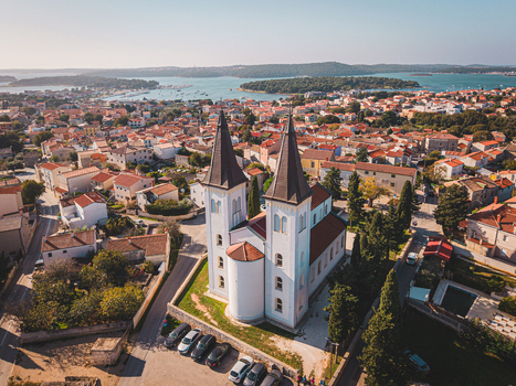 Aerial view of the towering church spires reach towards the sky contrasting against the terracotta rooftops and the shimmering Adriatic Sea Medulin Istria County Croatia. Aerial view of the towering church spires reach towards the sky contrasting against the terracotta rooftops and the shimmering Adriatic Sea Medulin Istria County Croatia.