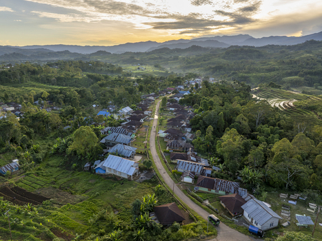 Aerial view of Bangka Kenda village with houses lining a road surrounded by lush green hills and rice terraces under golden hour light in Wae Rii East Nusa Tenggara Indonesia.