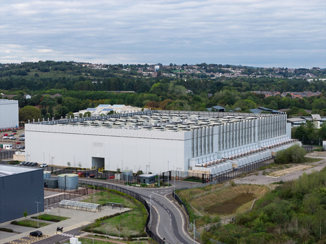 Aerial view of a large data center facility with industrial cooling units on the roof surrounded by green landscape and access roads in the United Kingdom.