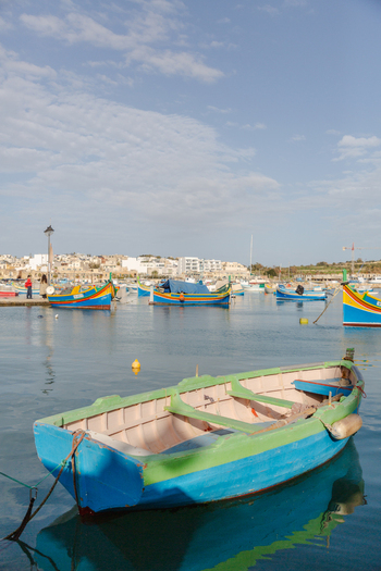 View of vibrant traditional fishing boats bob gently in the calm harbor waters reflecting the clear sky as the town basks in the warm sunlight Marsaxlokk Malta. View of vibrant traditional fishing boats bob gently in the calm harbor waters reflecting the clear sky as the town basks in the warm sunlight Marsaxlokk Malta.