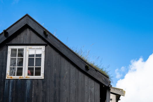 View of a traditional black wooden house with a white framed window and a lush green turf roof under a clear blue sky in the Faroe Islands. View of a traditional black wooden house with a white framed window and a lush green turf roof under a clear blue sky in the Faroe Islands.