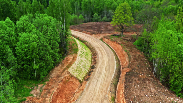 Aerial view of a winding dirt road cutting through a dense green forest with exposed red clay embankments in Latvia. Aerial view of a winding dirt road cutting through a dense green forest with exposed red clay embankments in Latvia.
