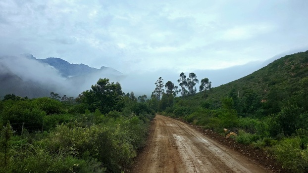 View of a gravel road winding through the Klein Karoo landscape near Towerkop with mist clinging to the mountains under an overcast sky in Ladismith Western Cape South Africa. View of a gravel road winding through the Klein Karoo landscape near Towerkop with mist clinging to the mountains under an overcast sky in Ladismith Western Cape South Africa.