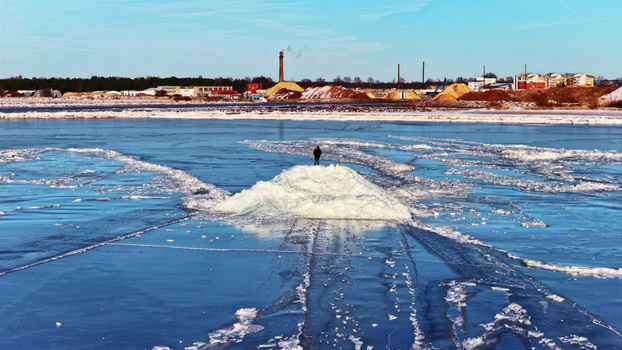 Aerial view of a person standing on a large ice mound in a frozen river with ice floes industrial buildings and wood chip piles in the background Latvia. Aerial view of a person standing on a large ice mound in a frozen river with ice floes industrial buildings and wood chip piles in the background Latvia.